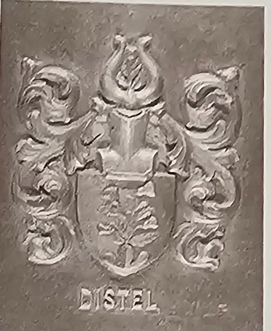 A carved relief plaque of the Distel arms, photographed in black and white: a thistle on a shield, a helm and mantling above it, and the name DISTEL carved below